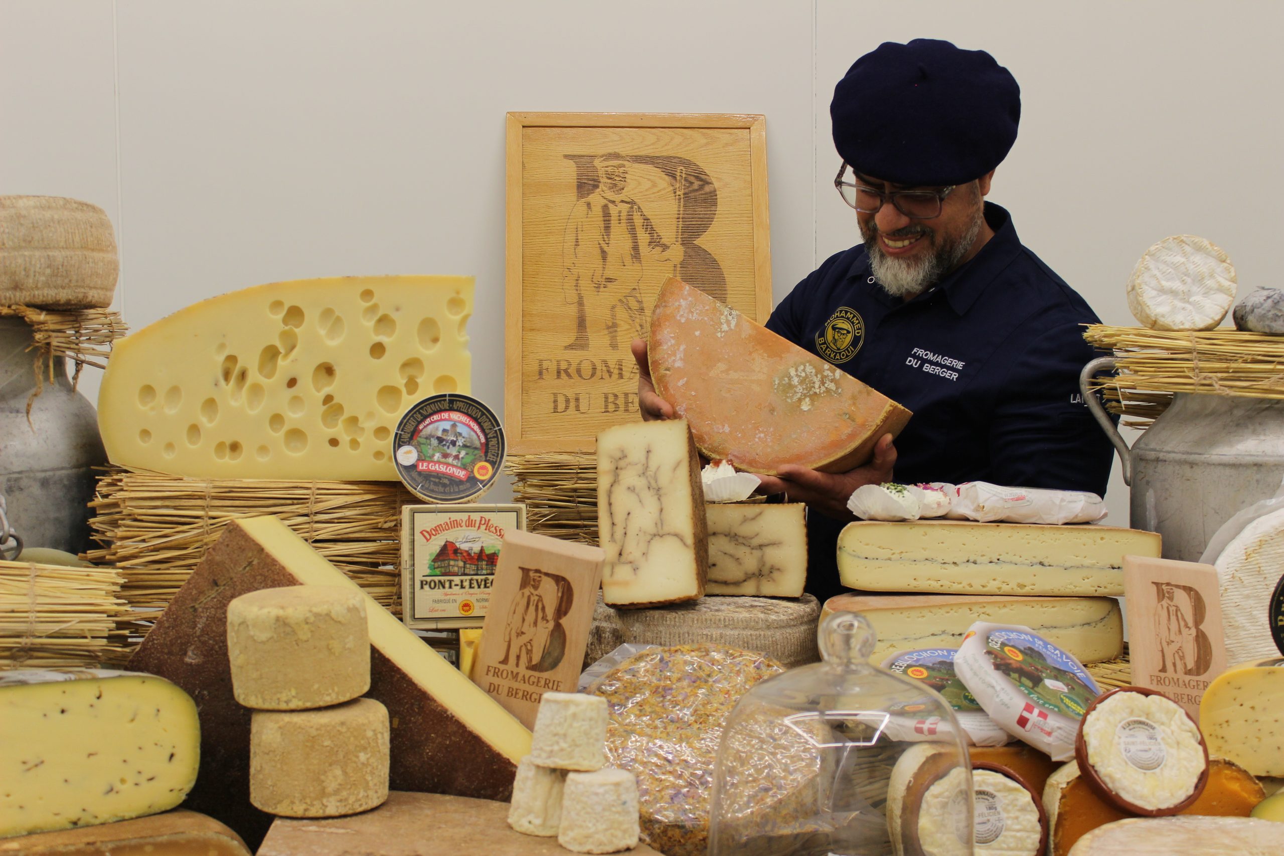 Mohammed Barkaoui, en béret et veste de la Fromagerie du Berger, présentant une demi-meule derrière un étal garni de fromages